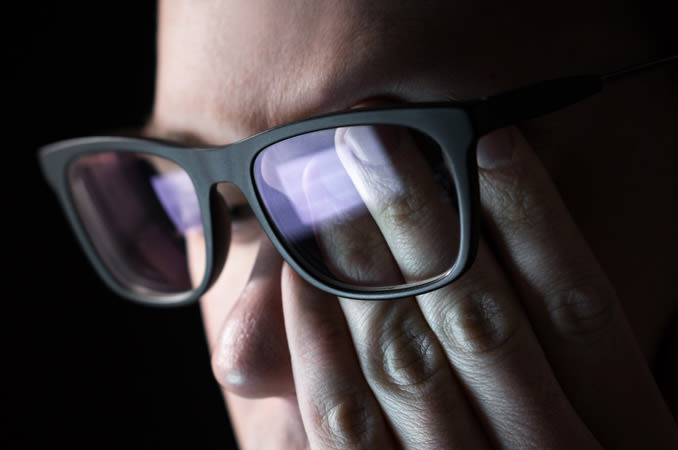 Man wearing glasses rubs his eye while looking at a computer.